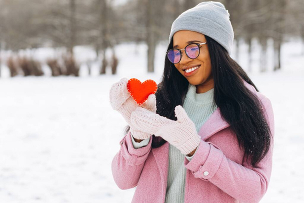 Young afro american woman with glasses and warm mittens holding a decorative red heart on Valentine's Day in winter. - Foto, imagen