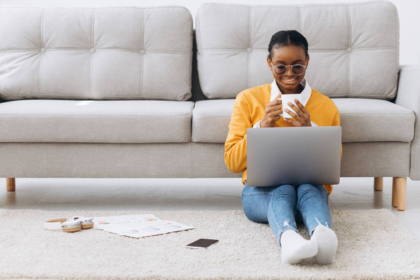 Beautiful young Black female university student sitting on the floor, drinking coffee and doing project on laptop - Photo, Image