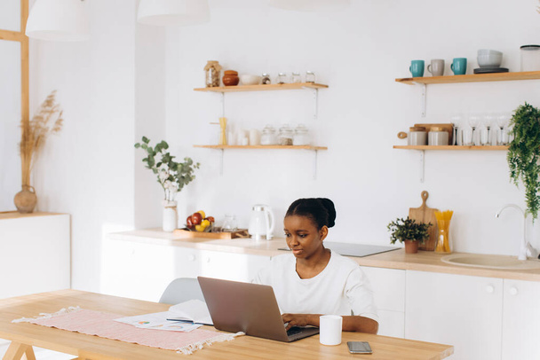 Retrato de uma jovem negra sentada na cozinha e trabalhando em um laptop de casa.  - Foto, Imagem