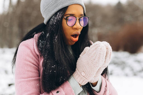 幸せな肯定的な女の子の肖像画、眼鏡や手袋でアフリカ系アメリカ人の若い女性は暖かい服で寒い霜の日に雪の中の冬の雪の公園で笑っています。冬のコンセプト - 写真・画像