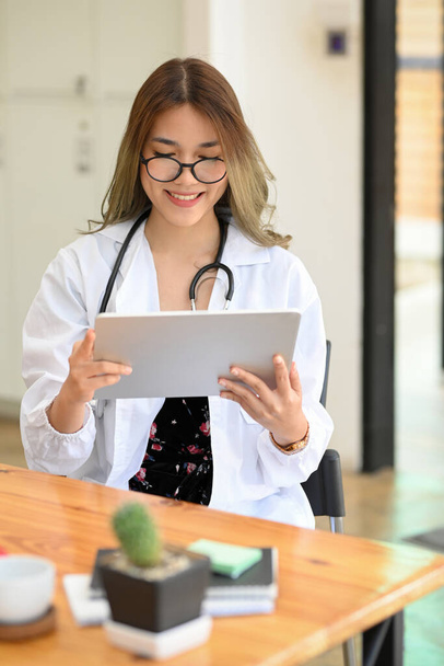 Retrato, lindo asiático feminino médico sentado em seu escritório mesa e verificação de histórico médico relatório no computador tablet digital. - Foto, Imagem