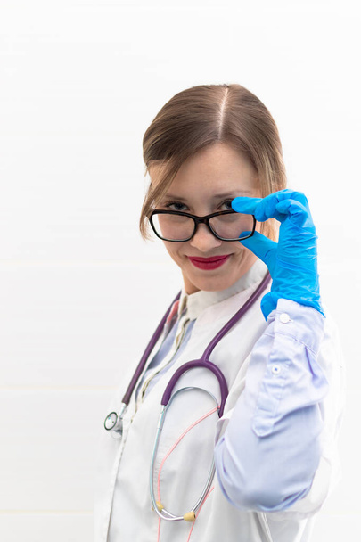 Mooie jonge arts vrouw in medische handschoenen en bril met een stethoscoop in een ziekenhuis op een witte achtergrond. Selectieve focus. Portret. Close-up - Foto, afbeelding