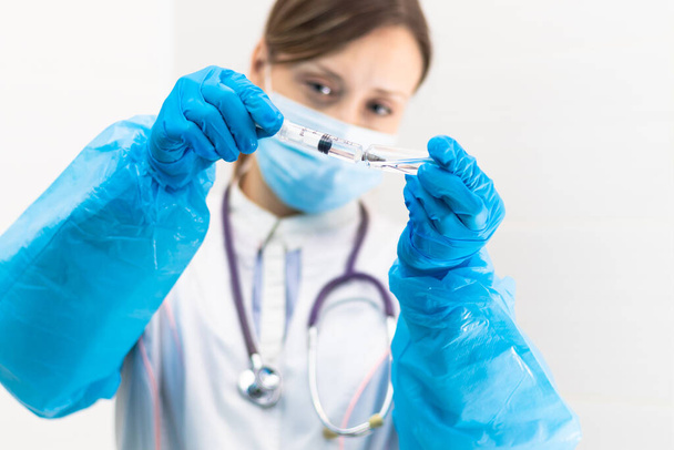 Jonge vrouwelijke arts met een medisch masker en handschoenen met een spuit in haar hand en een stethoscoop in een ziekenhuis tijdens een coronaviruspandemie. Selectieve focus. Portret - Foto, afbeelding