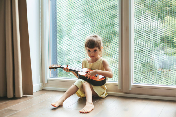 Portré egy aranyos kislány 4-5 éves játszik ukulele, ül a nagy ablak mellett. Gyönyörű gyermek tanul hangszeren játszani. - Fotó, kép