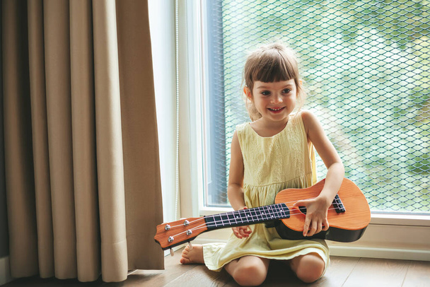 Ukulele tutan sevimli küçük kız, büyük pencerenin yanında oturuyor. Güzel bir çocuk müzik aleti çalmayı öğreniyor.. - Fotoğraf, Görsel