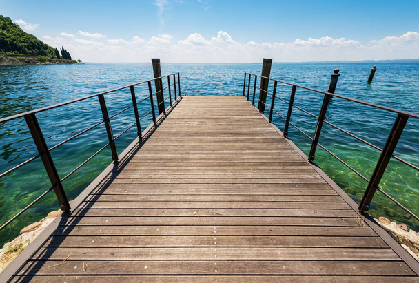 Małe puste drewniane molo na wybrzeżu jeziora Garda (Lago di Garda) przed małym miasteczkiem Garda, kurort turystyczny w prowincji Werona, Wenecja Euganejska, Włochy, Europa Południowa. - Zdjęcie, obraz