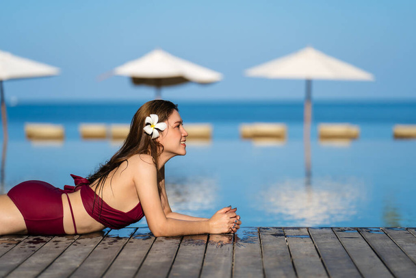 young woman in swimsuit relaxing on wooden edge of swimming pool with sea background - Фото, изображение