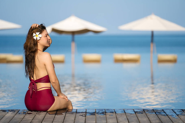 young woman in swimsuit sitting on wooden edge of swimming pool with sea background - Foto, afbeelding