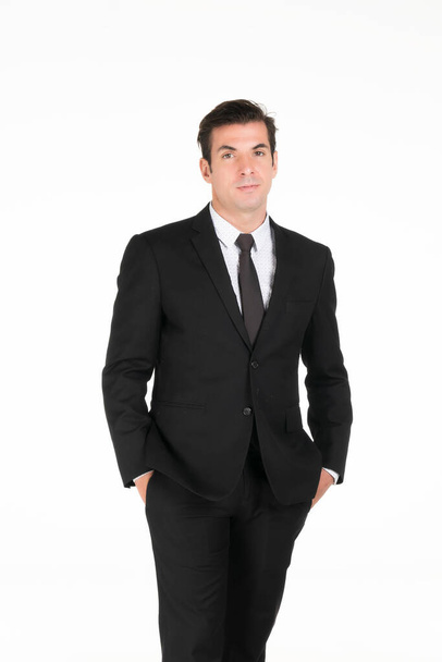 Portrait d'un homme d'affaires occidental souriant portant un costume noir les mains dans la poche de son pantalon au studio tourné sur fond blanc. - Photo, image