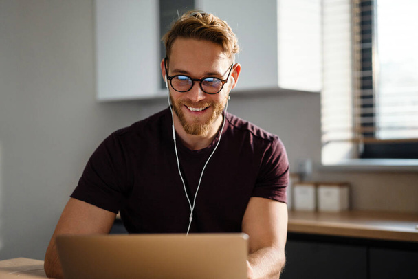 Young attractive man working remotely and heaving a meeting on a laptop while wearing earphones - Foto, Imagen