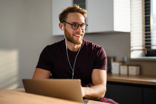 Young attractive man working remotely and heaving a meeting on a laptop while wearing earphones - Foto, Imagem