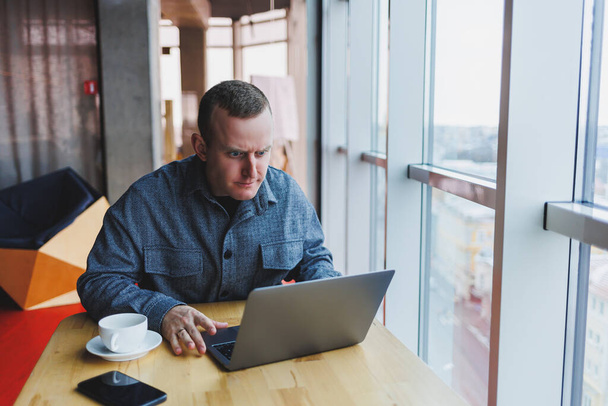 Επιτυχημένος επιχειρηματίας κάθεται σε ένα τραπέζι σε ένα καφέ, κρατώντας ένα φλιτζάνι καφέ και χρησιμοποιώντας ένα φορητό υπολογιστή. - Φωτογραφία, εικόνα