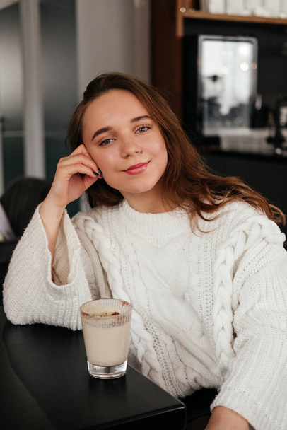 Jovencita sonriente bebiendo café sentada en la cafetería. Caucásico chica en suéter mirando a la cámara. - Foto, imagen