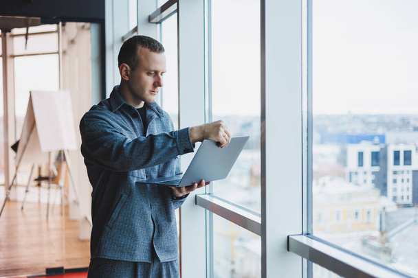 Retrato de um gerente inteligente inteligente do sexo masculino, ele está segurando um laptop para pesquisa no escritório, um homem de negócios em roupas elegantes jogando no touchpad. Em pé no escritório por uma grande janela - Foto, Imagem
