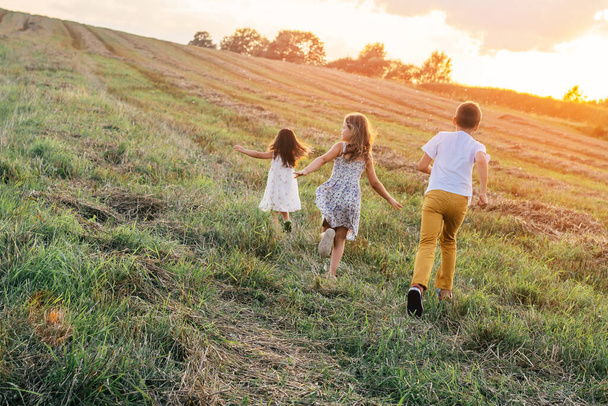 Portrait of three children playing game of catch and running on grass hay field paths of dry grass in the sunrise. - Photo, Image
