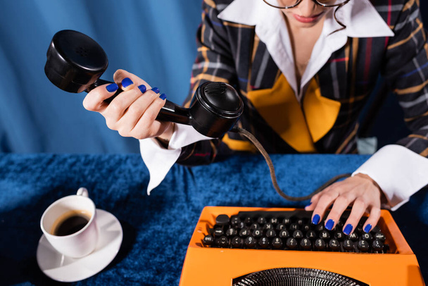 partial view of stylish newswoman holding handset while typing on vintage typewriter on blue background - 写真・画像