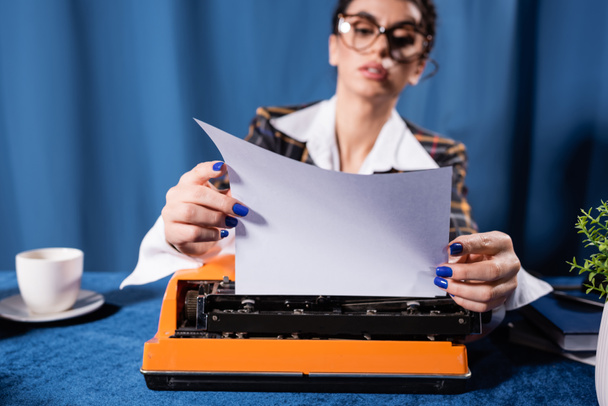 zamazana kobieta w okularach trzymająca pusty papier w pobliżu maszyny do pisania na niebieskim tle - Zdjęcie, obraz