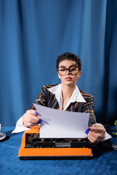 novinář ve stylu vintage oblečení drží prázdný papír v blízkosti psacího stroje na modrém pozadí - Fotografie, Obrázek