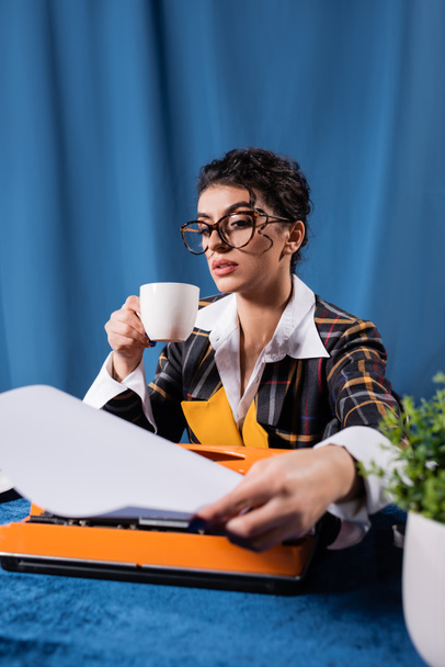 vintage style newswoman with coffee cup holding empty paper near typewriter on blue background - Photo, Image