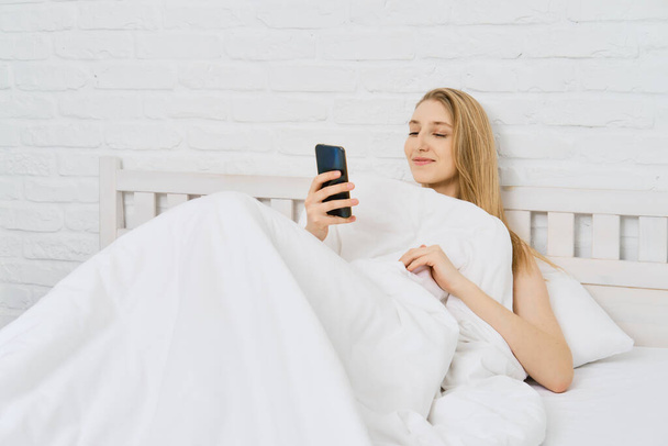 Young cheerful woman woke up, took smartphone and reading feed of social media - Foto, Imagen