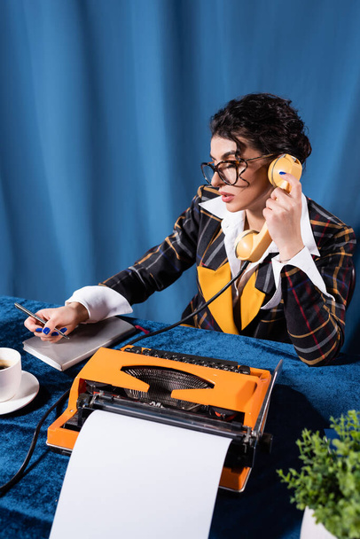brunetka newswoman w vintage marynarka trzymając długopis podczas rozmowy telefonicznej na niebieskim tle - Zdjęcie, obraz