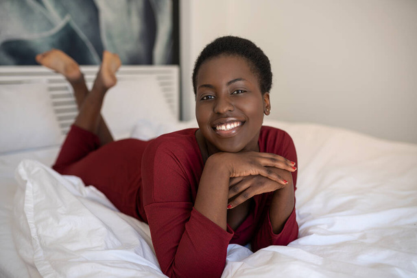 Πορτρέτο της όμορφης Μαύρης Αφρικανικής γυναίκας ξαπλωμένη στο κρεβάτι με τα πόδια και τα χέρια σταυρωμένα, χαμογελώντας και κοιτάζοντας στην κάμερα - Φωτογραφία, εικόνα