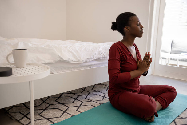 寝室のヨガマットに座って美しい黒アフリカの女性。瞑想とヨガの練習。早朝の運動。コピースペースで撮影 - 写真・画像