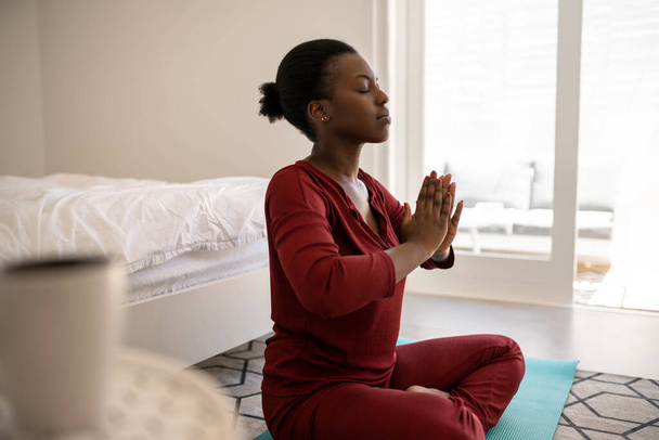 Gyönyörű fekete afrikai nő ül jóga szőnyegen a hálószoba padlóján. Meditál kéz a kézben és a szem csukva - Fotó, kép