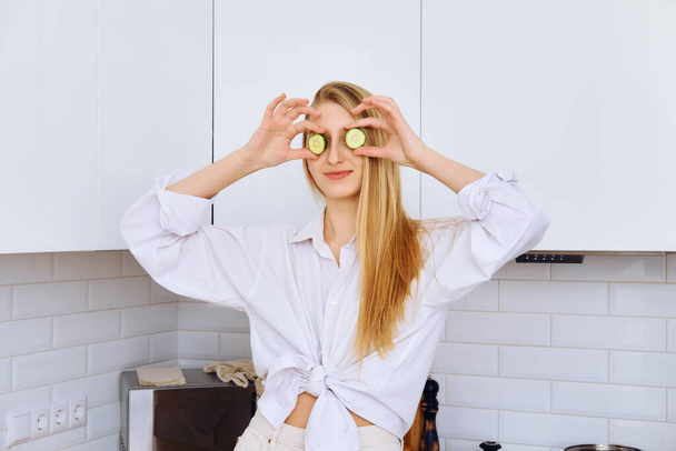 Joyful young woman put pieces of cucumber to her eyes while cooking - Fotografie, Obrázek