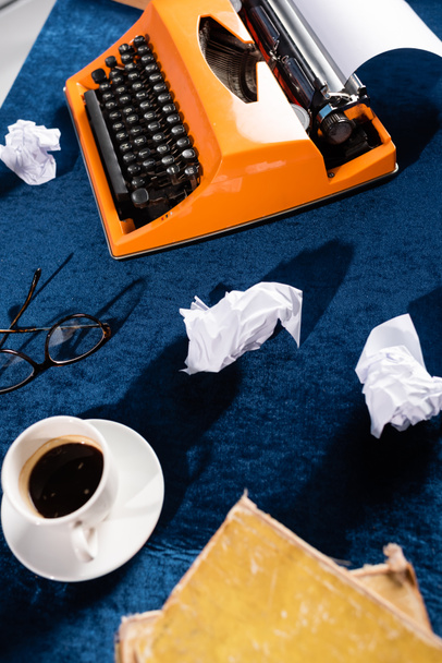 Draufsicht auf Kaffeetasse in der Nähe von Vintage-Schreibmaschine, Brille und zerknülltem Papier auf blauer Velours-Tischdecke - Foto, Bild