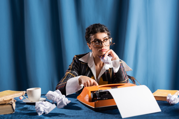 Nachdenkliche Zeitungsfrau, die neben zerknülltem Papier und alter Schreibmaschine auf blauem Hintergrund wegschaut - Foto, Bild