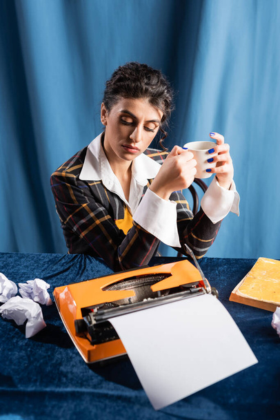 Journalist im Retro-Stil mit Kaffeetasse neben Schreibmaschine und zerknülltem Papier auf blauem Hintergrund - Foto, Bild