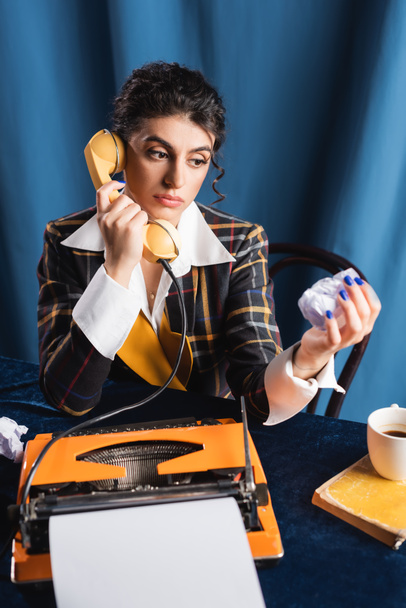 pensive nieuwsvrouw met verkreukeld papier praten over de telefoon in de buurt typemachine op blauwe achtergrond - Foto, afbeelding