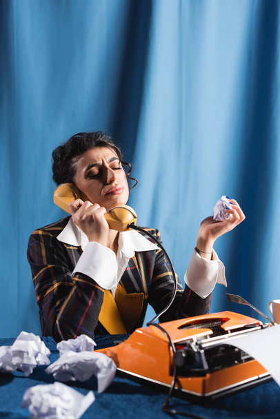 Mavi arka planda daktilonun yanında telefonla konuşurken buruşmuş kağıt tutan üzgün bir kadın. - Fotoğraf, Görsel