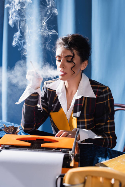 estilo vintage newswoman mecanografía en la máquina de escribir mientras se fuma sobre fondo azul - Foto, Imagen