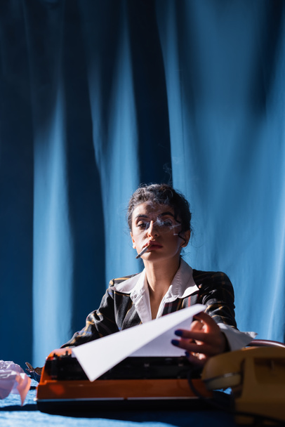 Курение винтажный стиль женщина держит бумагу рядом с пишущей машинкой на синем фоне с драпировкой - Фото, изображение