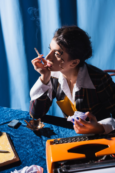 periodista reflexivo con cigarrillo y papel arrugado sentado cerca de la máquina de escribir vintage sobre fondo azul - Foto, Imagen