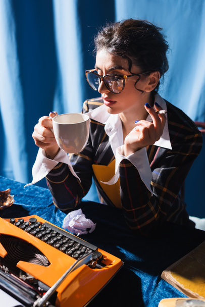 vintage styl newswoman v brýlích kouření a pití kávy v blízkosti psacího stroje na modrém pozadí - Fotografie, Obrázek
