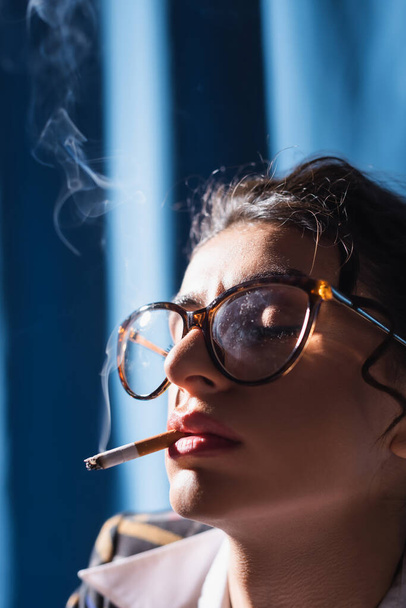 close up view of woman in vintage eyeglasses smoking with closed eyes on blue background - Fotoğraf, Görsel