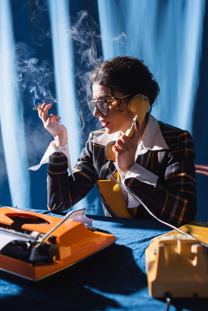 periodista de estilo retro con cigarrillo hablando por teléfono cerca de la máquina de escribir sobre fondo azul - Foto, Imagen