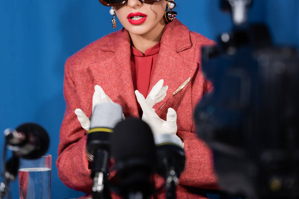 blurred microphones near cropped woman in white gloves giving interview on blue background - Zdjęcie, obraz