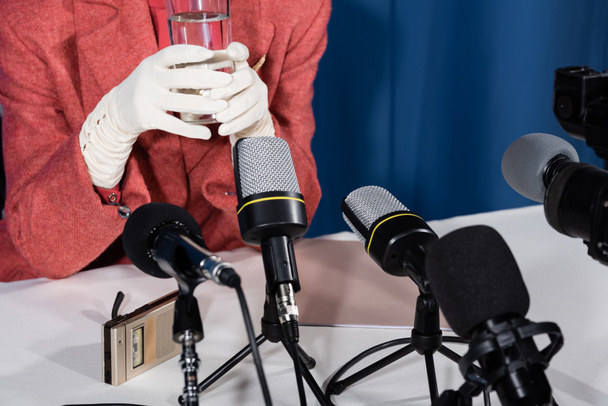 vista cortada de mulher em luvas brancas segurando vidro de água perto de microfones e gravador de voz no fundo azul - Foto, Imagem