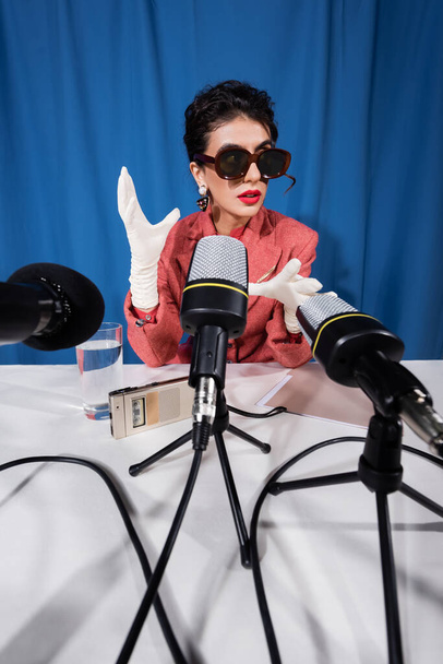 mikrofonok közelében vintage stílusú nő napszemüvegben gesztikuláló interjú során kék háttér - Fotó, kép