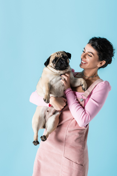 young and cheerful woman cuddling pug dog while standing isolated on blue - Φωτογραφία, εικόνα