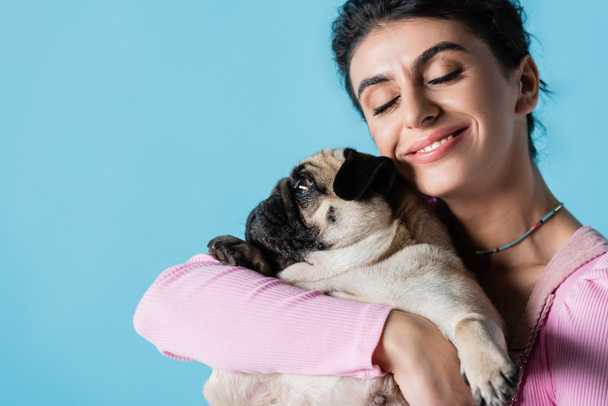 happy brunette woman with closed eyes hugging pug dog isolated on blue - Fotó, kép