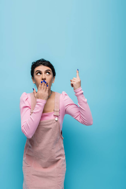 shocked woman looking up, pointing with finger and covering mouth with hand isolated on blue - Foto, Imagem
