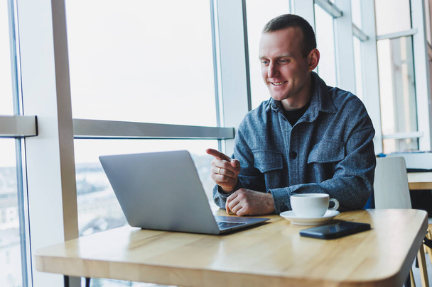 Homem de negócios feliz bem sucedido está sentado a uma mesa em um café, segurando uma xícara de café e usando um laptop. - Foto, Imagem