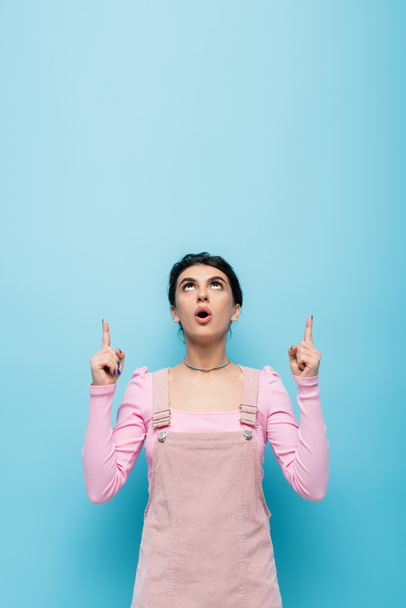 astonished woman in pastel clothing looking up and pointing with fingers isolated on blue - Фото, изображение