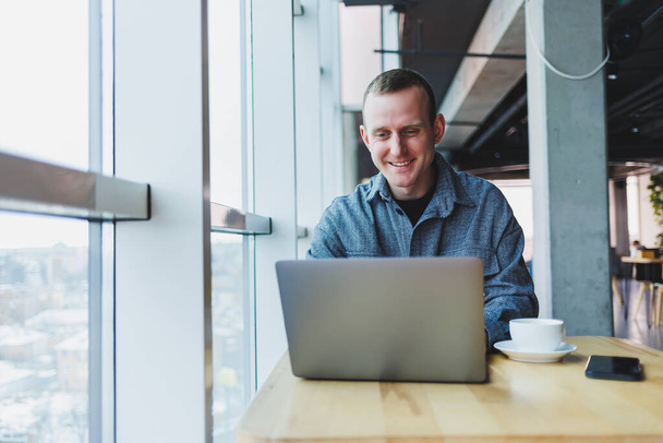 Homem de negócios feliz bem sucedido está sentado a uma mesa em um café, segurando uma xícara de café e usando um laptop. - Foto, Imagem