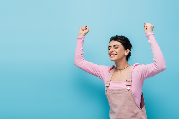 cheerful and stylish woman with closed eyes showing triumph gesture on blue background - Foto, afbeelding